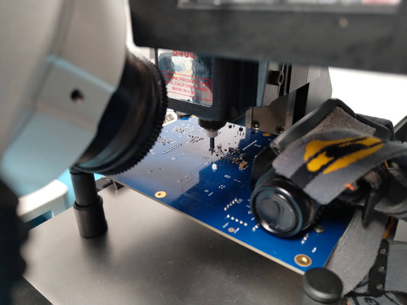Blue PCB mounted upside down on the table of a milling machine with a microscope pointed at it